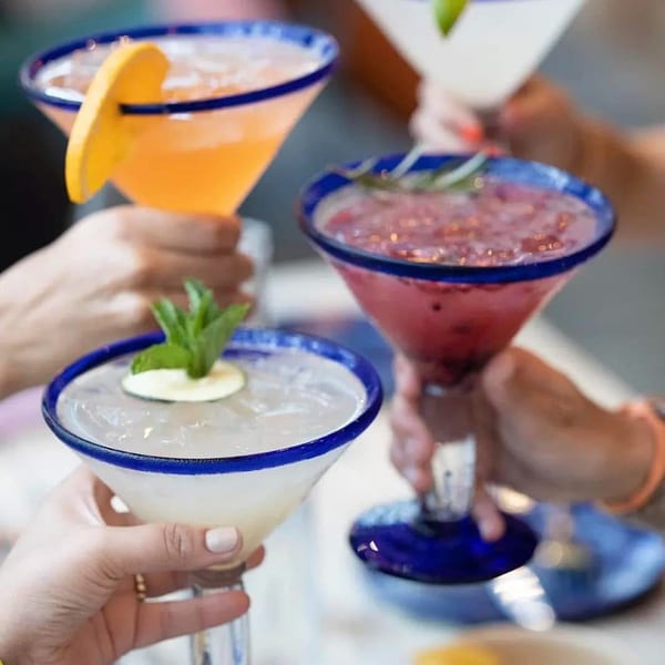 Four hands hold up colorful margaritas in blue-rimmed glasses, garnished with fruit and mint.