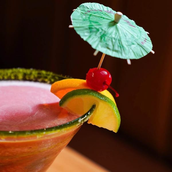Pink margarita with a green sugar rim, garnished with lime, orange, cherry, and a paper umbrella.