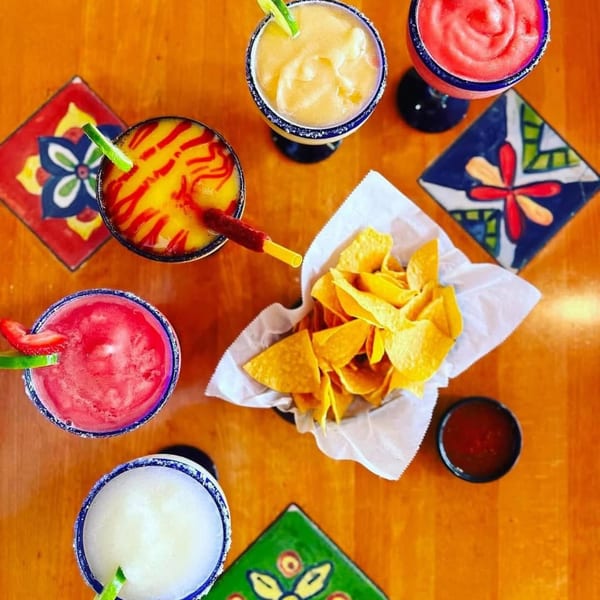 Overhead view of five colorful margaritas, a basket of tortilla chips, and salsa on a wooden table.