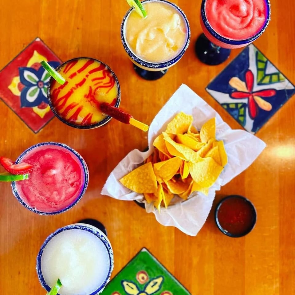 Overhead view of five colorful margaritas, a basket of tortilla chips, and salsa on a wooden table.
