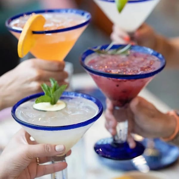 Four hands holding colorful margaritas with blue-rimmed glasses, garnished with fruit and mint.