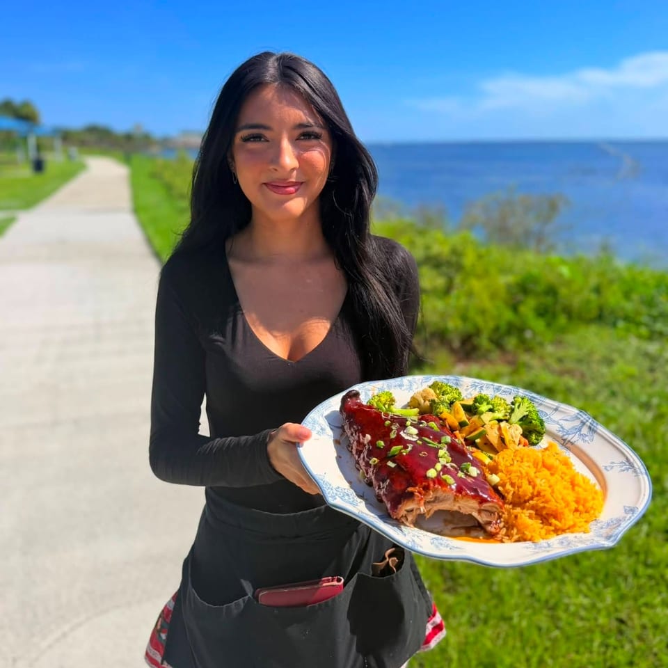 Woman holding a plate of glazed ribs, orange rice, roasted vegetables, and broccoli beside a sunny oceanfront path.