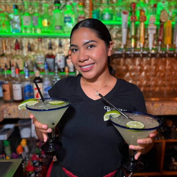 A smiling Chaparral server holds two large margaritas with lime slices and straws in a bar setting.
