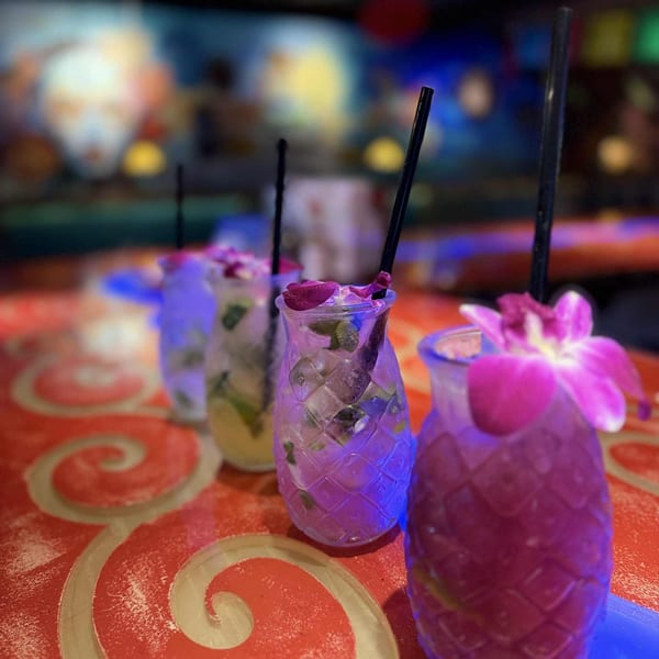 Four colorful pineapple-shaped cocktails with straws and floral garnishes on a red patterned table.