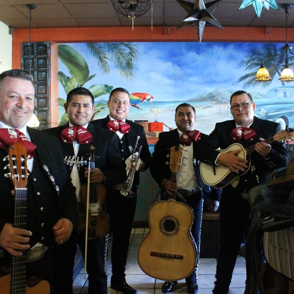 Five smiling mariachi musicians in black suits and red bow ties, holding instruments, in a restaurant.
