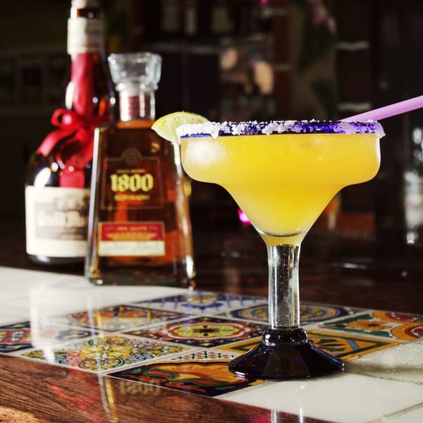 A vibrant yellow margarita with a salted rim and lime wedge sits on a colorful tiled bar.