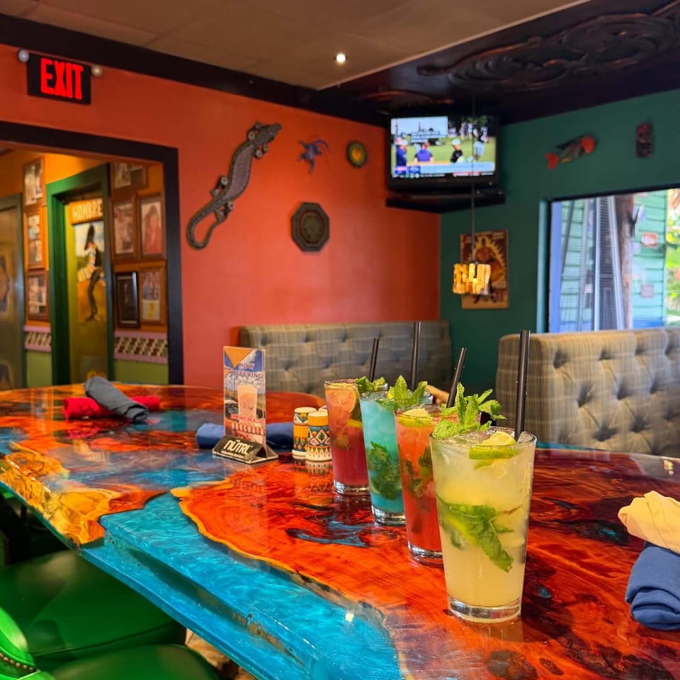 Colorful restaurant booth with four garnished cocktails on a vibrant painted table, surrounded by eclectic decor and a television.