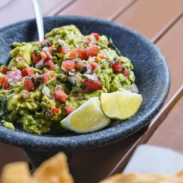 Fresh guacamole topped with diced tomatoes and onions, garnished with lime wedges in a dark bowl.