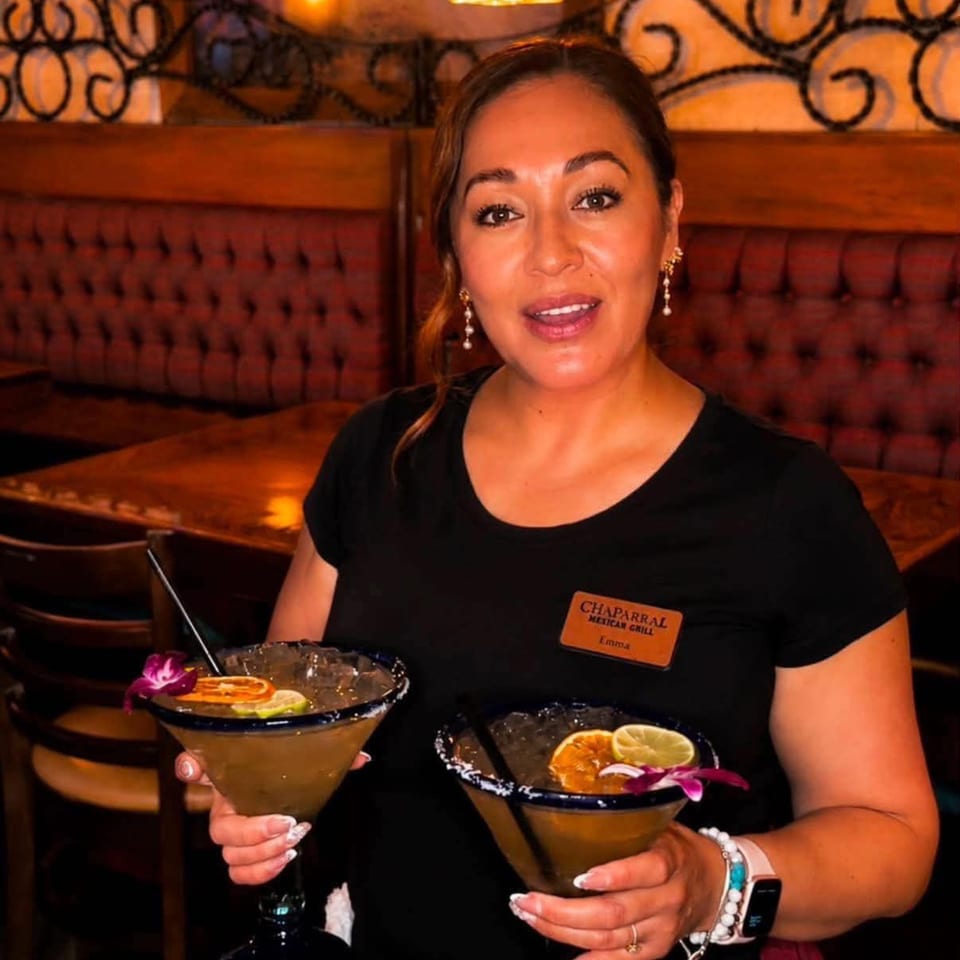 Restaurant server holding two garnished margaritas in a warmly lit dining room with leather booths.