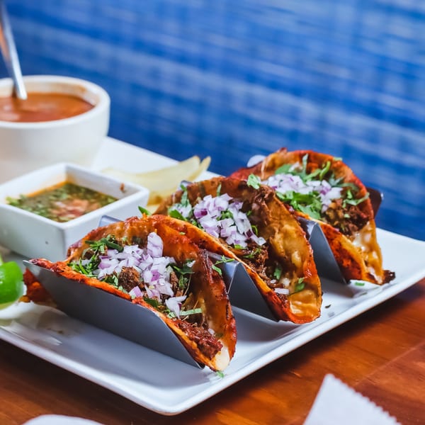 Three birria tacos with red onions and cilantro, served with consommé and green salsa.
