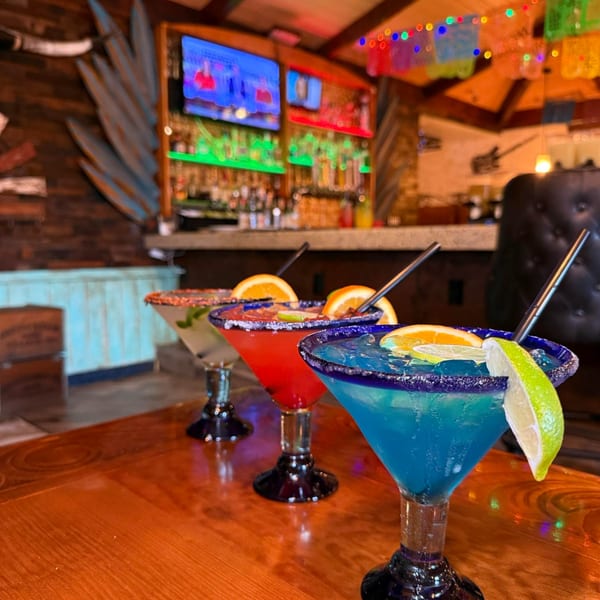 Three colorful margaritas with fruit garnishes on a wooden table in a vibrant Mexican restaurant.